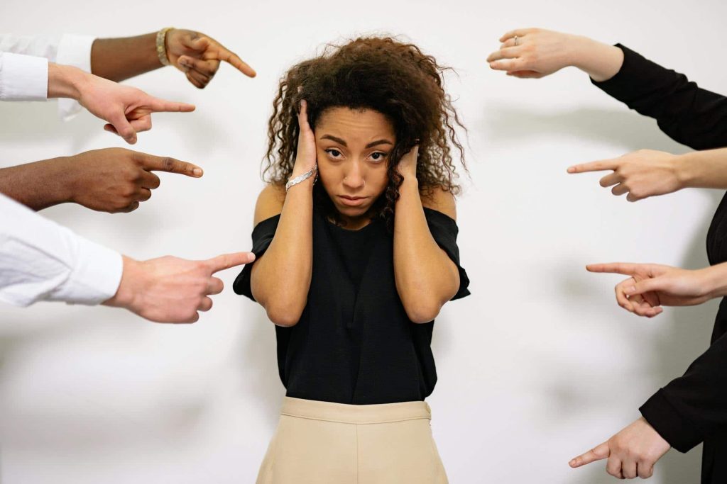 Woman with fingers from many people pointing at her, with hands to her ears.